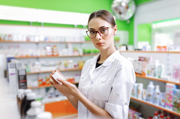 Uma jovem magra de cabelos castanhos, de óculos, vestida com um jaleco branco, olha para a câmera e segura um pequeno frasco no saguão de uma nova farmácia. .