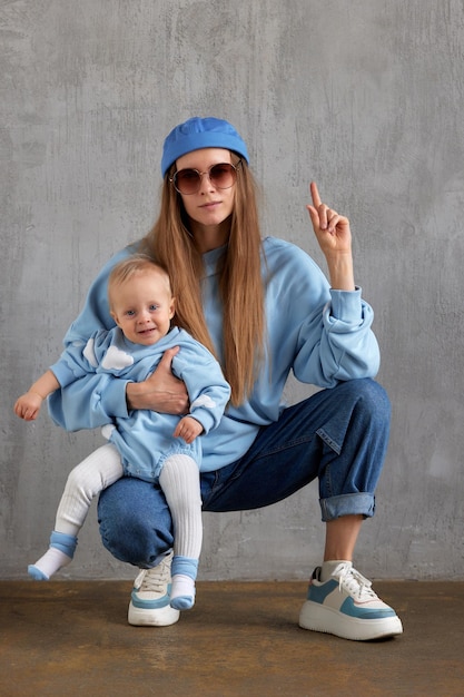 Uma jovem mamãe elegante em jeans, um suéter azul, um boné azul engraçado e óculos de sol sentados em seus quadris