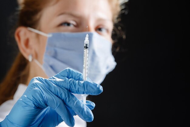 Uma jovem médica com uma seringa olha para a câmera uma enfermeira usando uma máscara médica e lat azul