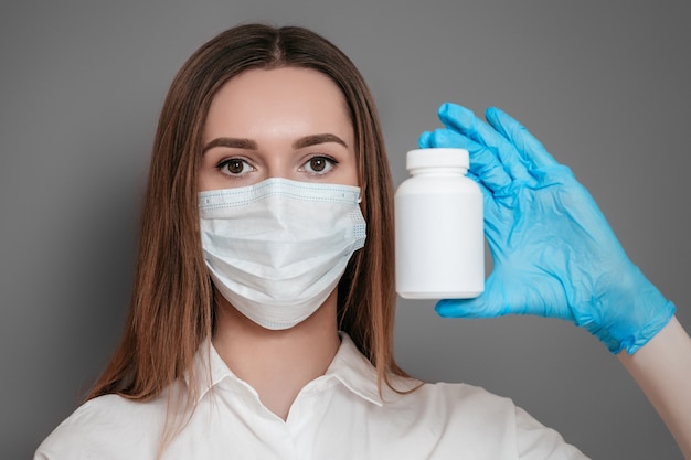 Uma jovem médica virologista em uma máscara médica protetora no rosto, segurando um frasco de comprimidos