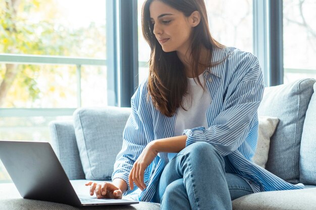 Uma jovem moderna escrevendo no computador sentada no sofá em um quarto de hotel, viagem ou apartamento interno em casa, lendo e escrevendo e-mails ou atividade de notificação on-line.