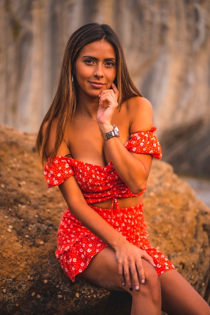 Uma jovem morena caucasiana em um vestido vermelho na praia de Itzurrun, na cidade de Zumaia, Guipúscoa. País Basco. Sessão de estilo de vida, retrato, sentado em uma pedra do mar verde com olhar sedutor