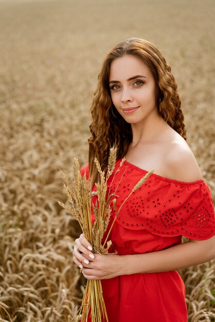 Uma jovem morena com um vestido de verão vermelho segura espigas de um campo de trigo. Liberdade.