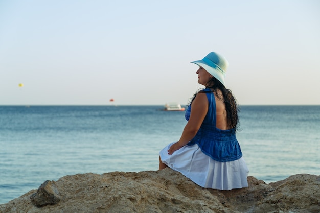 Uma jovem morena de saia branca e chapéu azul à beira-mar está sentada de costas