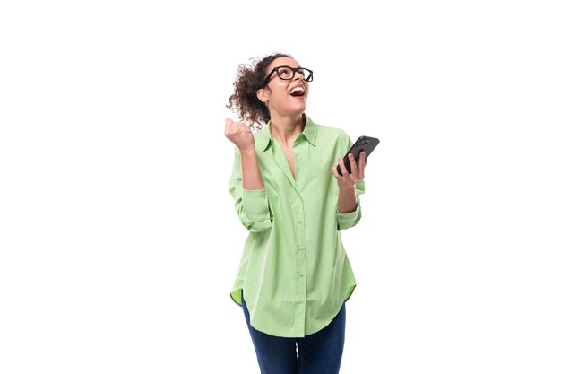 Foto uma jovem morena e encaracolada de óculos vestida com uma camisa verde está conversando em um smartphone