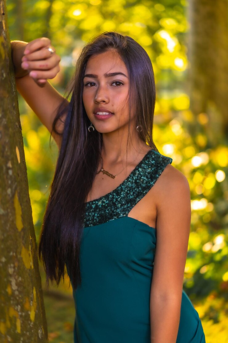 Foto uma jovem morena latina com cabelos longos e retos, encostado em uma árvore em um vestido verde