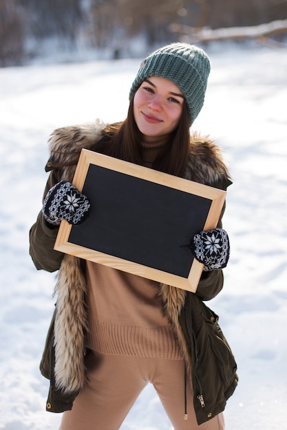 Uma jovem, morena, quadro em mãos, quadro-negro, espaço de cópia, contra o pano de fundo da paisagem de inverno. Neve e geada, o conceito de Natal.