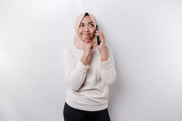 Uma jovem muçulmana asiática pensativa usando um hijab e segurando o queixo enquanto fala ao telefone isolada por um fundo branco
