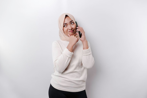 Uma jovem muçulmana asiática pensativa usando um hijab e segurando o queixo enquanto fala ao telefone isolada por um fundo branco