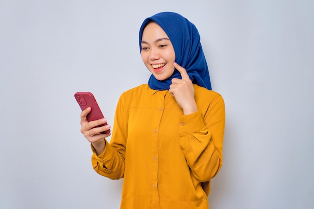 Uma jovem muçulmana asiática sorridente vestida de camisa laranja olhando para a tela do celular se sente alegre lendo boas notícias isoladas sobre fundo branco