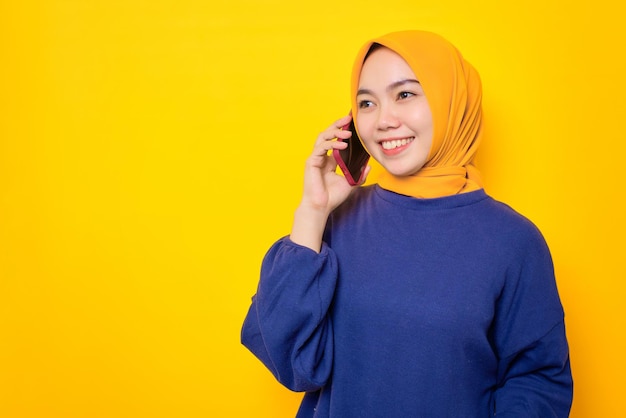 Uma jovem muçulmana asiática sorridente vestida de suéter casual falando no celular com espaço de cópia isolado sobre fundo amarelo