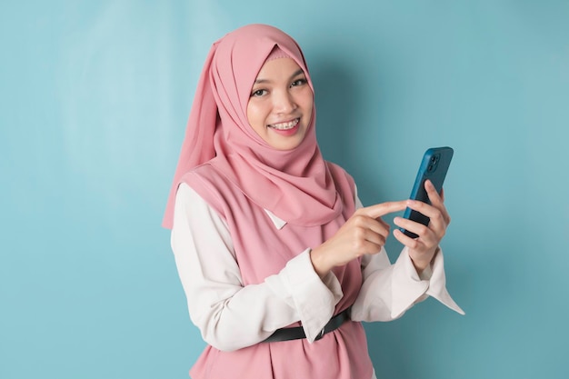 Uma jovem muçulmana asiática usando hijab rosa está sorrindo enquanto segura seu smartphone