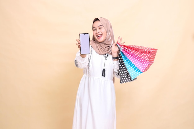 uma jovem muçulmana asiática usando um hijab sorri felizmente mostrando uma tela de celular e carregando um