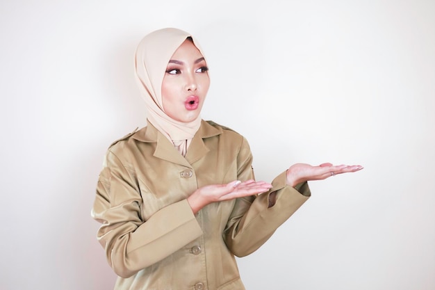 Uma jovem muçulmana asiática vestindo um uniforme marrom e hijab apontando para o espaço vazio no fundo isolado branco do estúdio