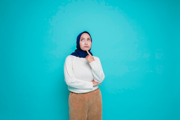Uma jovem muçulmana pensativa está vestindo camisa branca e hijab segurando o queixo enquanto olha de lado isolada por fundo azul