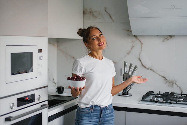 Uma jovem muito sorridente segura um prato de bagas frescas de cereja na mão na cozinha