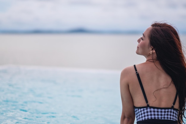 Uma jovem mulher asiática relaxando em uma piscina infinita olhando para uma bela vista do mar