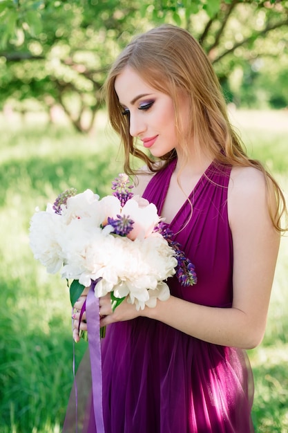 Uma jovem mulher atraente em um vestido roxo e com maquiagem profissional posa em um pomar.