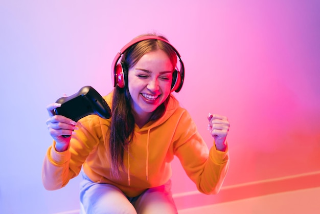 Uma jovem mulher bonita caucasiana com um capuz amarelo jogando um videogame usando um joystick e fones de ouvido com surpresa de foco divertido e uma cara de vitória vencedora com luz neon azul e vermelha