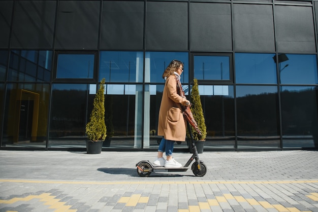 Uma jovem mulher bonita com um casaco a sorrir e a conduzir uma scooter eléctrica para o trabalho