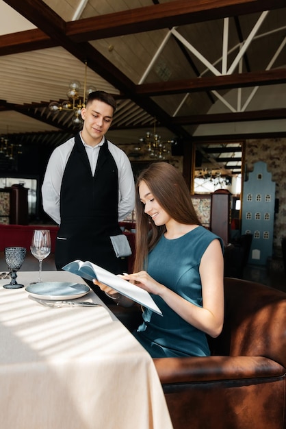 Uma jovem mulher bonita em um restaurante elegante examina o menu e faz um pedido para um jovem garçom em um avental elegante Atendimento ao cliente no restaurante e estabelecimentos de catering