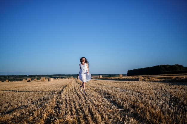 Uma jovem mulher bonita em um vestido branco de verão fica em um campo de trigo ceifado com enormes feixes de feno, curtindo a natureza. Natureza na aldeia. Foco seletivo