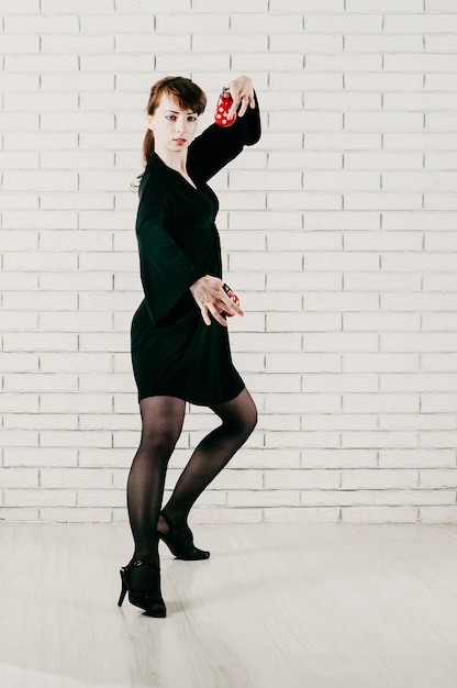 Foto uma jovem mulher bonita em um vestido preto dançando flamenco com castanholas vermelhas e fundo de tijolos brancos