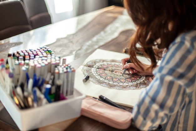 Uma jovem mulher bonita está sentada na cozinha de casa e desenhando um desenho colorido de mandala zen