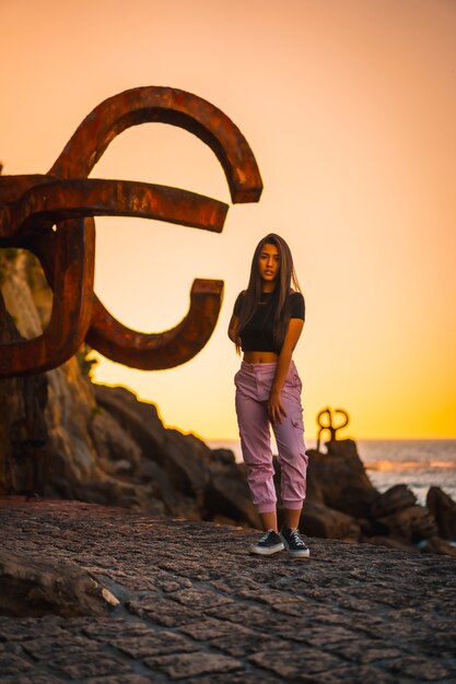 Uma jovem mulher bonita morena com cabelos longos e lisos em uma camiseta preta curta ao lado de uma famosa escultura de San Sebatian chamada Peine del Viento