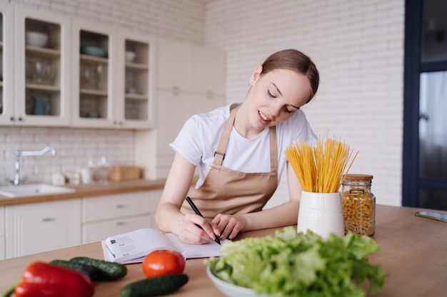 Uma jovem mulher bonita na cozinha, de avental, escreve suas receitas favoritas ao lado de legumes frescos