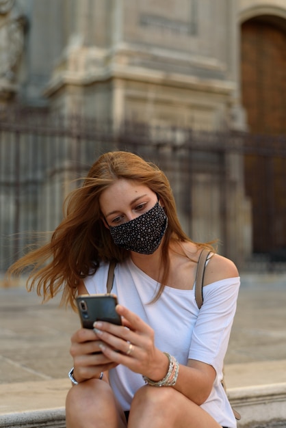 Foto uma jovem mulher branca com uma máscara olhando através de seu telefone em uma rua