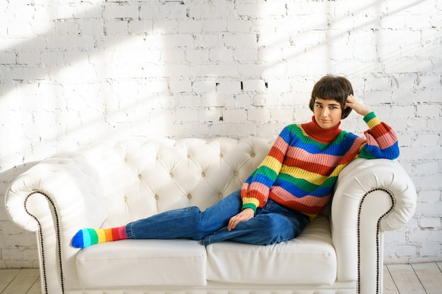Uma jovem mulher com cabelo curto em um suéter arco-íris e meias está sentada em um sofá branco, o conceito de minorias sexuais