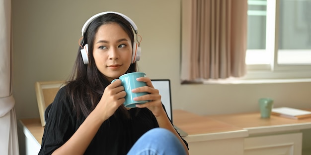 Uma jovem mulher com o fone de ouvido está segurando uma xícara de café enquanto está sentado e ouvindo música.