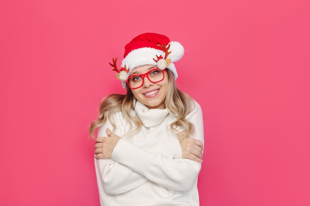 Uma jovem mulher com um suéter branco e óculos de Natal com chapéu de Papai Noel aprecia a suavidade abraçando a si mesma