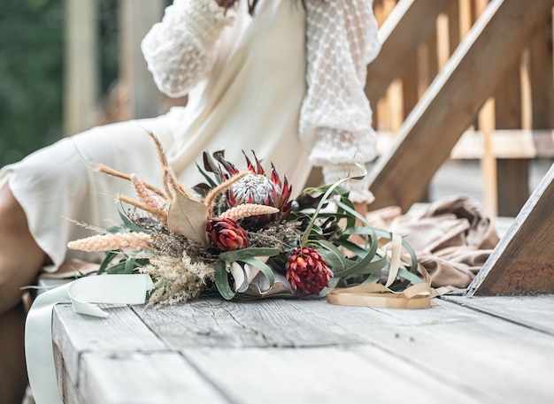 Uma jovem mulher com um vestido branco está sentada em uma ponte de madeira com um buquê de flores exóticas protea.