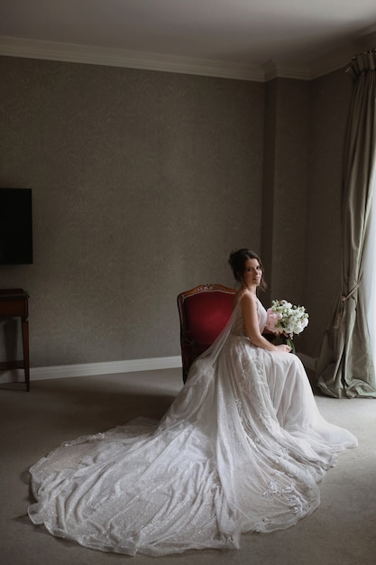 Uma jovem mulher com um vestido de noiva vintage com um buquê de flores está sentada na cadeira no interior vintage