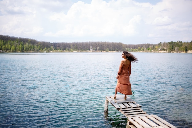 Uma jovem mulher com um vestido fica em alvenaria de madeira no meio de um lago azul. menina feliz sorri e o sol brilha, dia de verão. ela tem cabelos longos e cacheados e uma aparência europeia