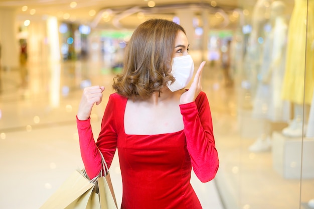 Uma jovem mulher com um vestido vermelho usando máscara protetora em shopping center, fazendo compras sob o conceito de pandemia de Covid-19.