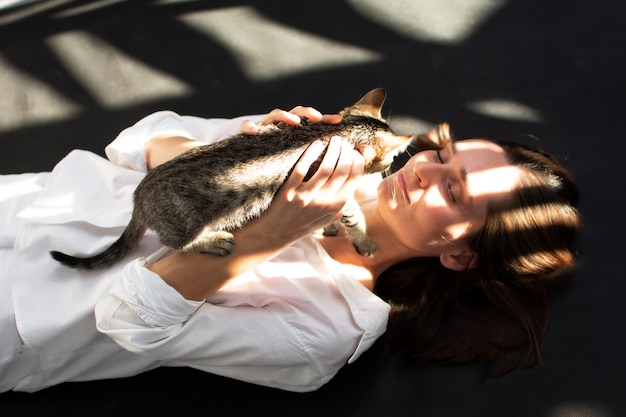 Uma jovem mulher com uma camisa branca está deitada e segurando um gatinho nos braços