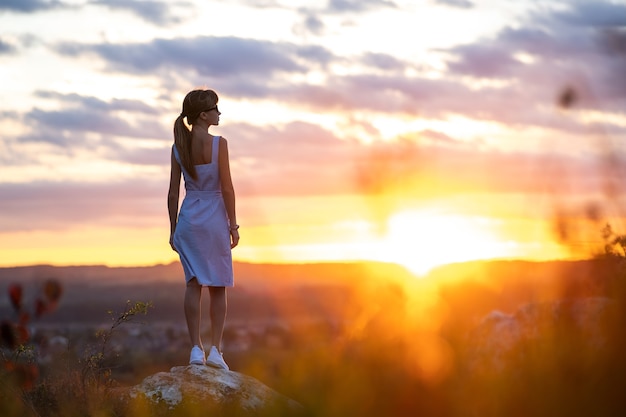 Uma jovem mulher com vestido de verão em pé ao ar livre, apreciando a vista do pôr do sol amarelo brilhante.