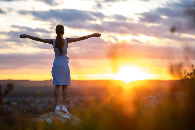 Uma jovem mulher com vestido de verão em pé ao ar livre com os braços estendidos, apreciando a vista do pôr do sol amarelo brilhante.