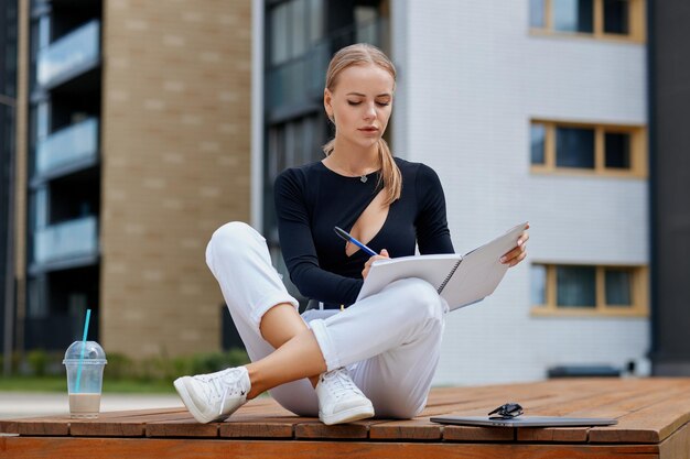 Uma jovem mulher de negócios com cabelos loiros vestida com estilo senta-se em um banco com um laptop em um campus de edifício residencial