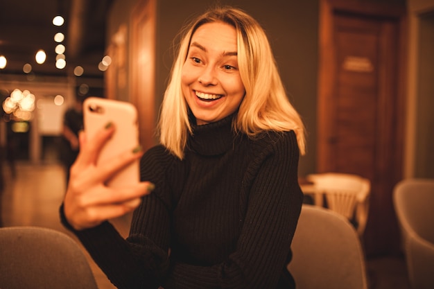 Uma jovem mulher está sentada em um café e fala ao telefone. Loira feliz está descansando em um café Foto de retrato de uma mulher em um suéter preto