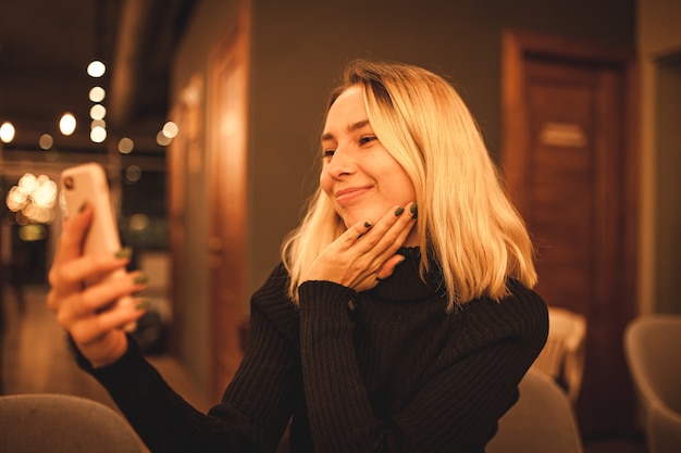 Uma jovem mulher está sentada em um café e fala ao telefone. Loira feliz está descansando em um café Foto de retrato de uma mulher em um suéter preto