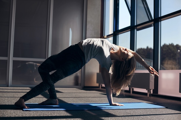 Uma jovem mulher faz yoga. A garota faz um asana com as costas dobradas, se apoia nas pernas e uma mão, a segunda mão é levantada. Vista lateral