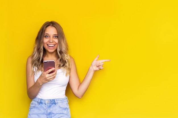Uma jovem mulher feliz aponta para copiar o espaço vazio para texto ou desenho com o dedo segurando um telefone celular