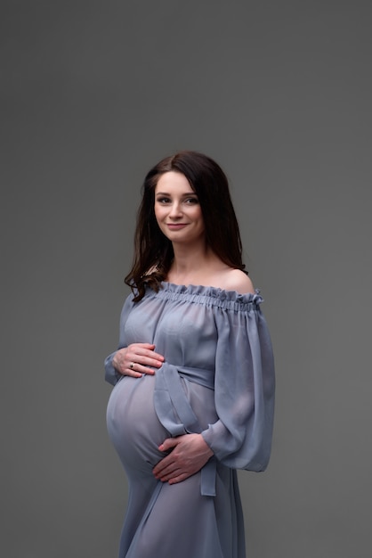 Uma jovem mulher grávida em um vestido fica em uma parede cinza