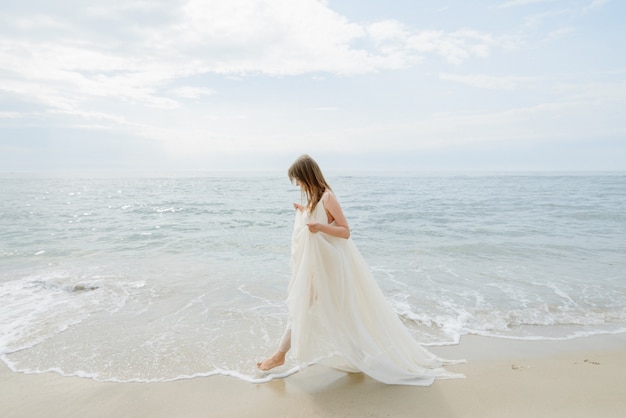 Uma jovem mulher linda em um vestido longo branco caminha ao longo da praia e cais contra o mar.