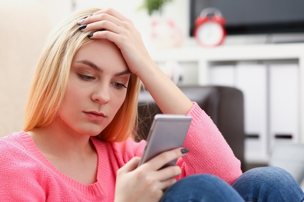 Uma jovem mulher loira e bonita sentada no sofá segurando o smartphone nas mãos