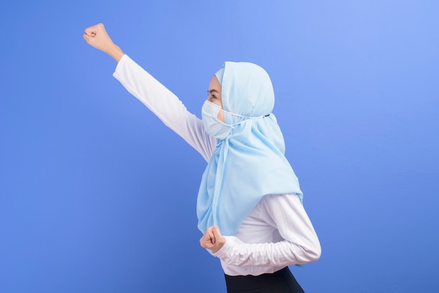 Uma jovem mulher muçulmana com hijab usando uma máscara cirúrgica sobre uma parede azul.